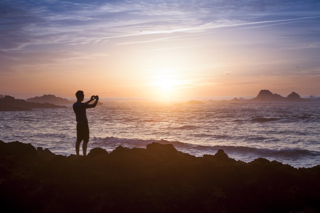 Man står på strand och fotar solnedgången med mobiltelefon.
