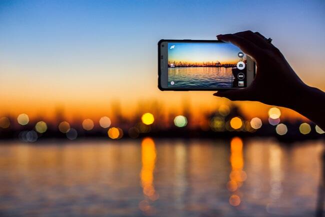 Hand fotograferar hamn i skymningen med mobiltelefon.